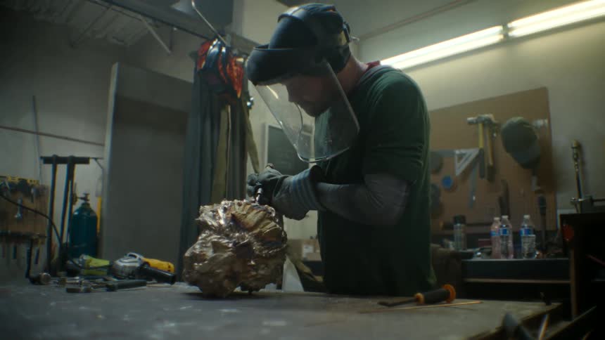 A metal sculptor using a drill to work on a metal sculpture of a head