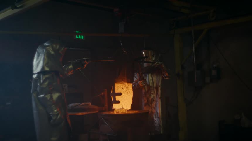 Two workers wearing fire proximity suits extract a redhot crucible from a forge