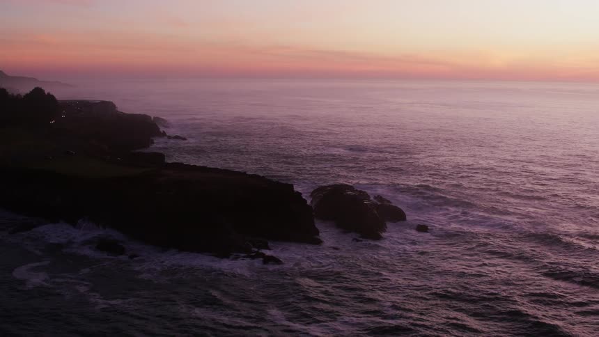 An aerial of ocean waves crashing against a rocky coast as the sun is setting