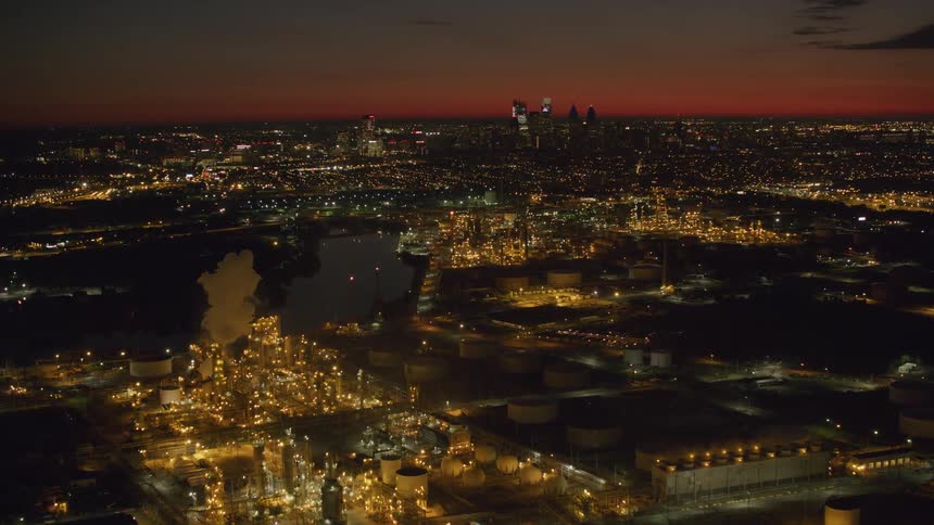 Refinery glows brightly a distance from downtown skyline under a red sky at night