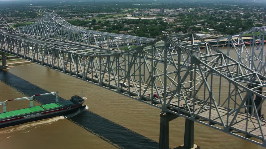 An aerial shot of a metal bridge over a city river with a long boat traveling underneath it