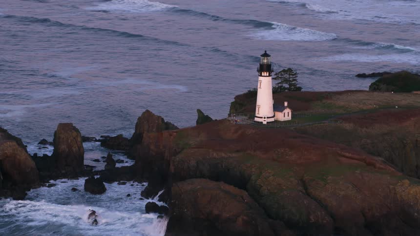 An aerial of a white lighthouse on the coast as waves from the ocean crash against the rocks
