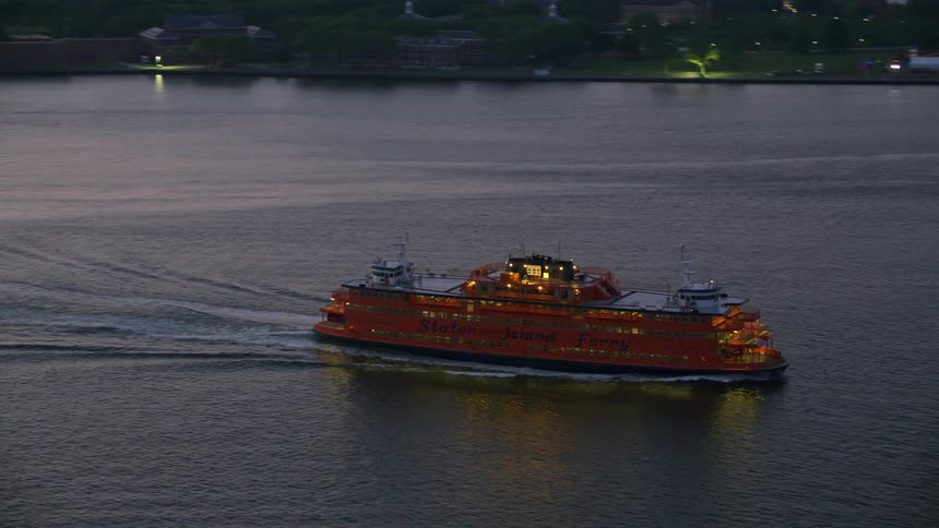 An aerial shot of a large ship moving through the waterway near a city at dusk