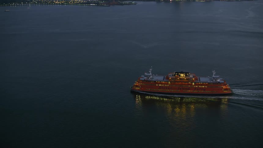 Drone view of a passenger ship lit up sailing across the waters with city skylines along the landscape horizon