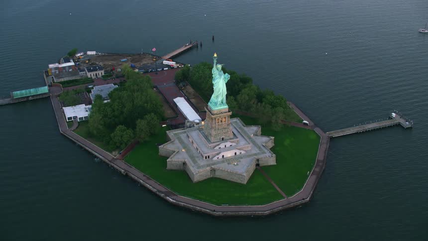 The statue of liberty stands on liberty island overlooking the hudson river holding her torch high in the air