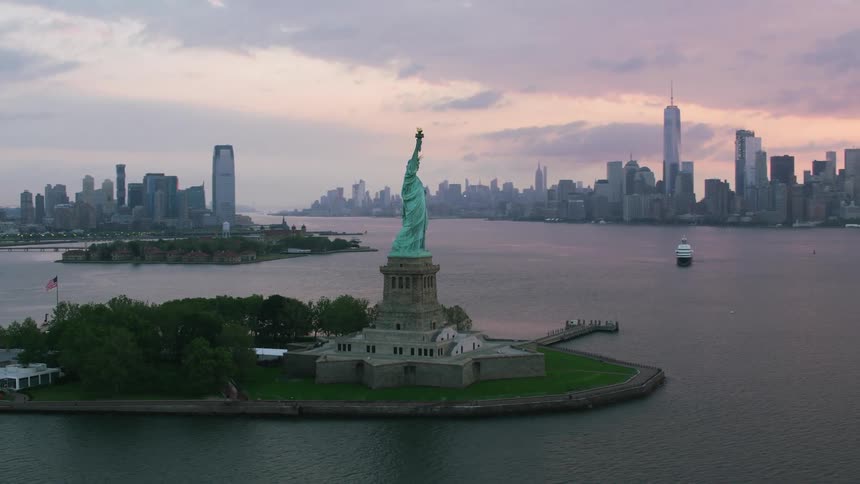 The statue of liberty stands tall on liberty island overlooking new york harbor as the sun sets behind buildings and towers on the city skyline