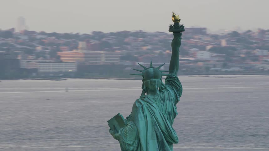 The statue of liberty stands tall overlooking the hudson river with holding her torch high in the air as boats sail in the water and city buildings are in the distance