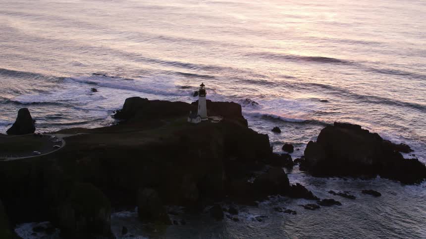 A lighthouse stands on a peninsula with cars parked nearby as waves roll in and crash against the rocks