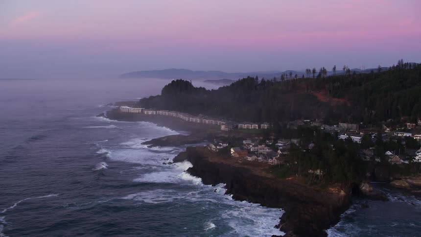 Waves roll onto coast strewn with apartment buildings on foggy day under a red tinted sky