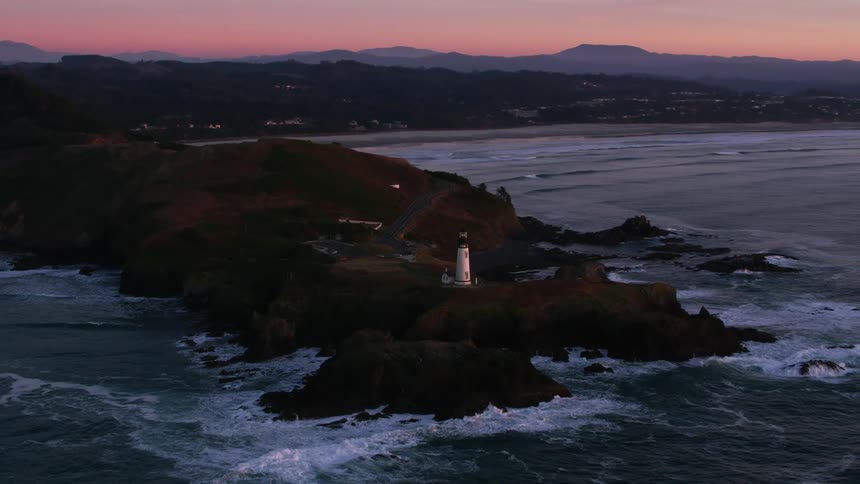 Ocean waves crashing against a rocky shore with a lighthouse at sunset