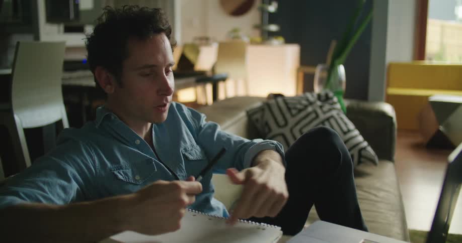 A man in a room sitting on a couch with a pen and a book and video chatting on a laptop