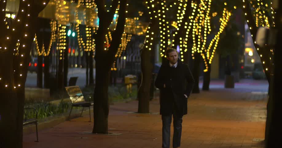 A well-dressed man walks alone on a city sidewalk lined with trees garnished with string lights