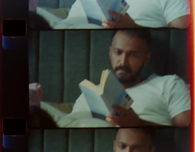 A man is sitting up in bed reading a book as a woman is lying across the bed reading