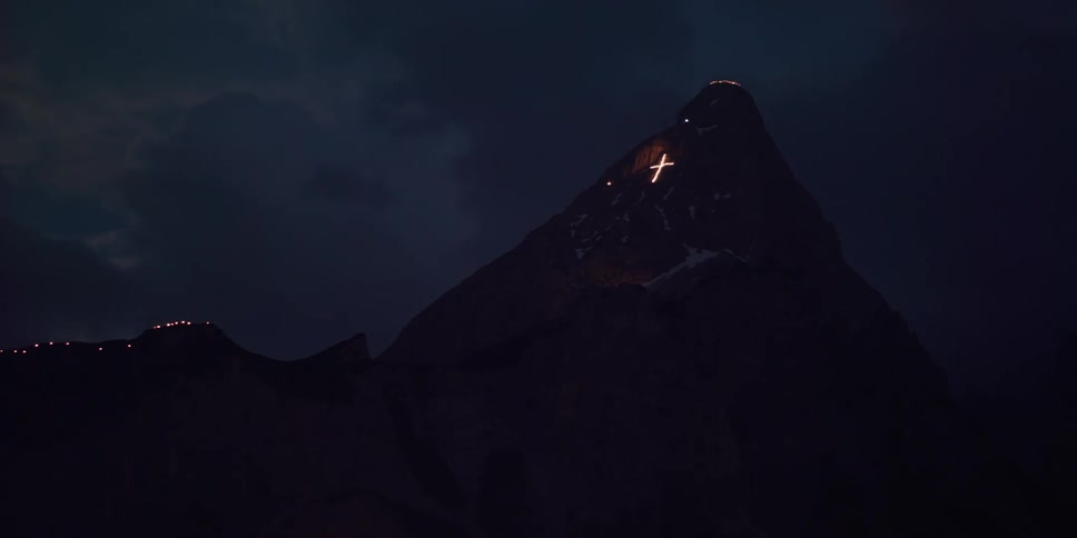 Fire lights glow across a mountain ridge and on a high mountain peak as a luminous cross shines on the mountainside in the darkness