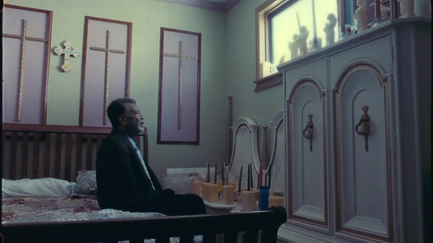 An older man sitting on the edge of his bed in front of candles while he looks out the window above him