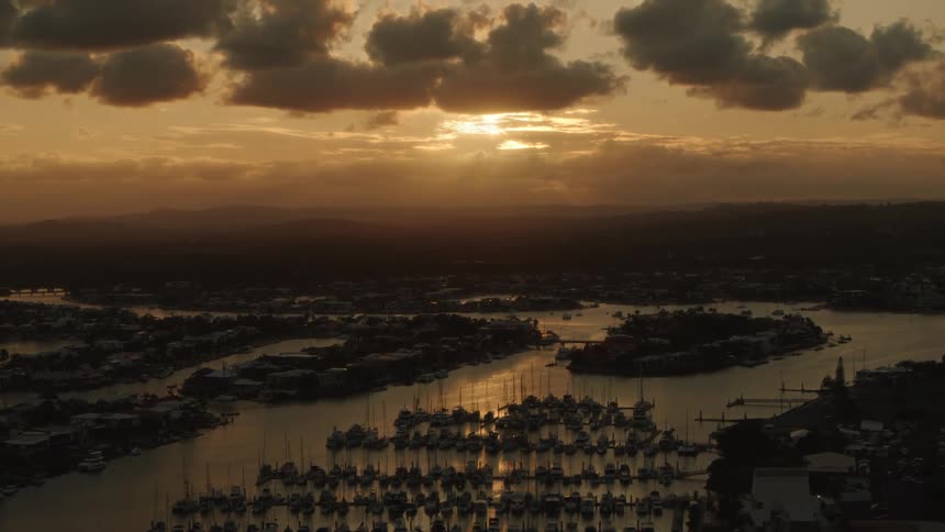 Boats are docked in a marina of a coastal community as the sun sets in a cloudy sky