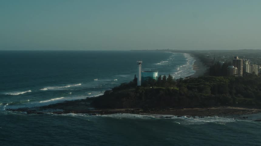 An aerial of a coastal city with waves crashing against the beach with a tree line along the coast