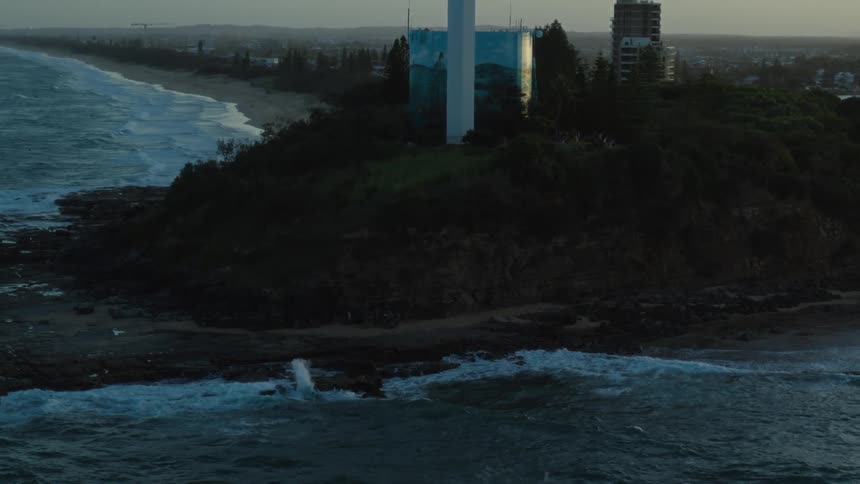 Waves roll into shore and splash along the coast as a tower next to a building overlooks the water