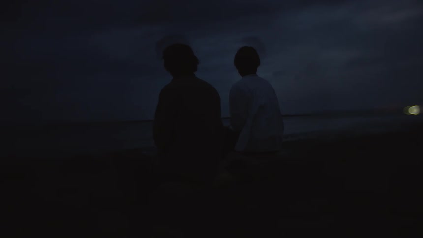 Two people sit together on beach looking out to the water at night