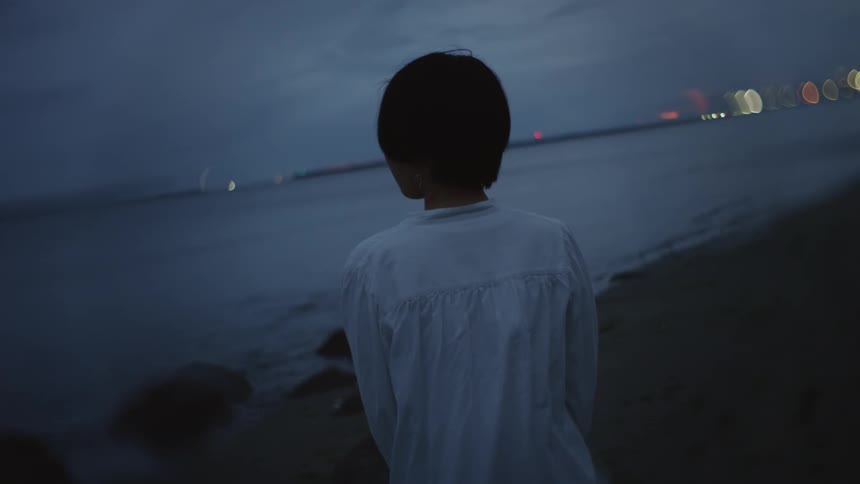Young woman wearing a white blouse standing on the beach at sundown staring across the sea