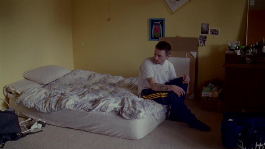 A man sitting on the edge of his mattress that sits on the floor and looks back and forward thinking