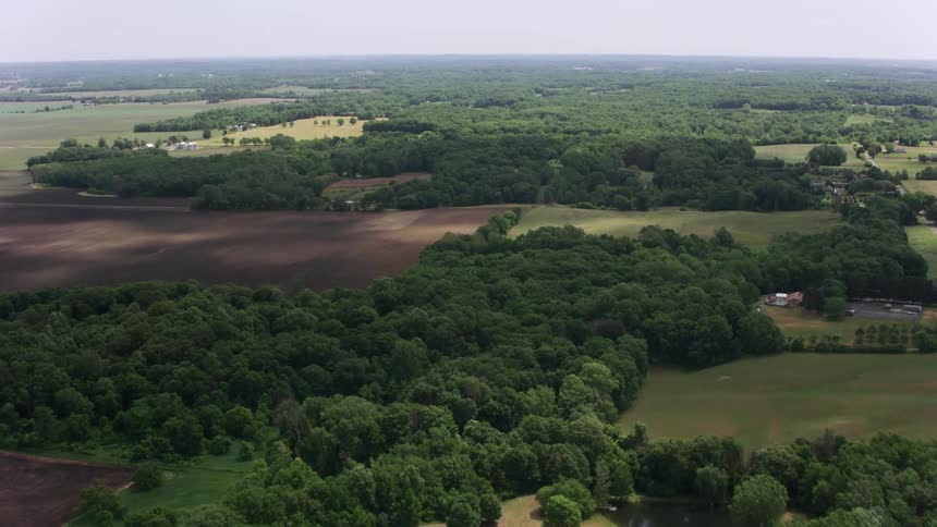 An automobile travels on a country road with houses far apart separated by trees and woods with open fields