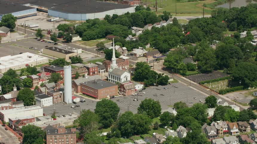 A town church with a large pointed tower and a large parking lot surrounded by trees and houses