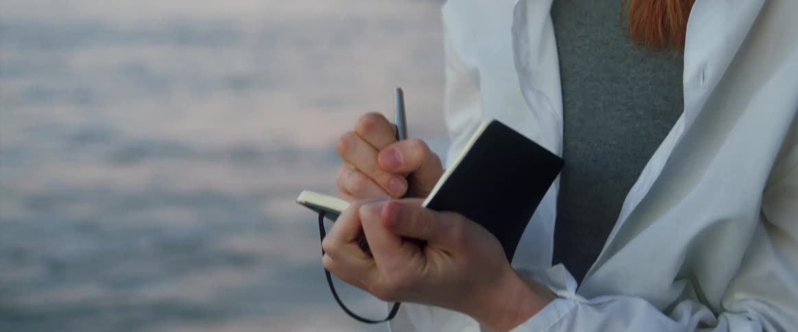A woman with a pen and a pad writing on a beach with a fence behind her