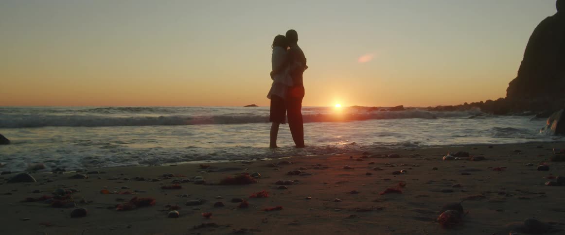A man wraps a sweater around a woman as they stand close together on the beach