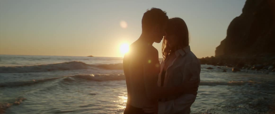 A man and woman embrace and look in each others eyes as they stand on the beach next to the ocean