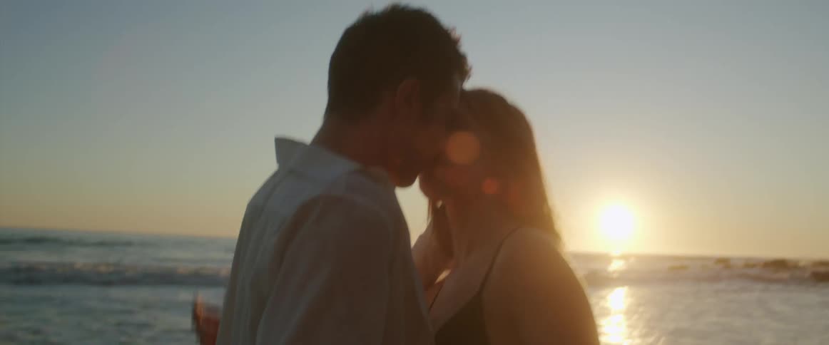 A young couple embraces and kisses on a beach as the sun sets behind them
