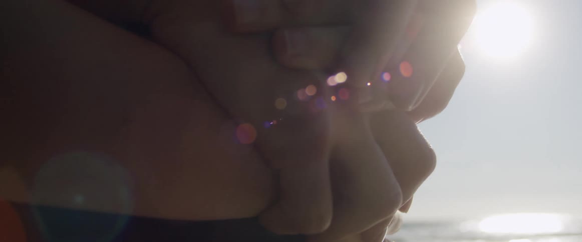 Man hugs woman from behind and they entwine hands while standing on beach