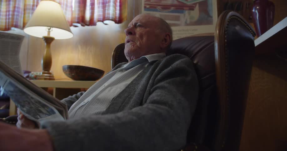Elderly man with gray cardigan shirt and jeans sits in brown armchair reading newspaper shaking his head