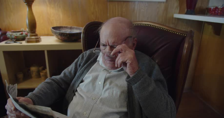 Elderly man sitting in a chair taking his glasses off while looking at a newspaper before putting it down on his lap and shaking his head