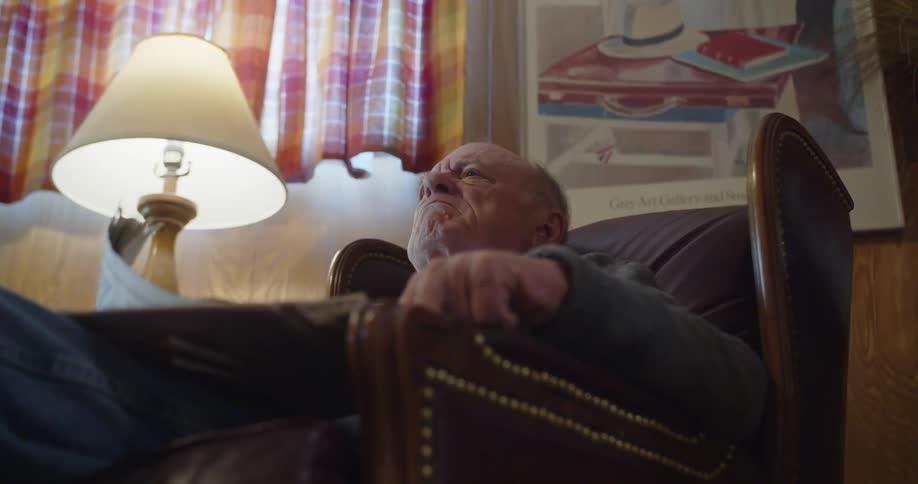 Elderly man sitting in brown armchair reads newspaper shaking his head