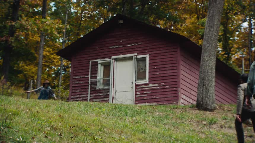 Two people walk up to a red cabin in the woods to find two teenagers running around it