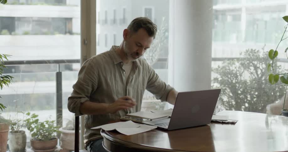 Man sitting at a round table in a room with windows all around and potted plants while he looks at papers and his computer in front of him