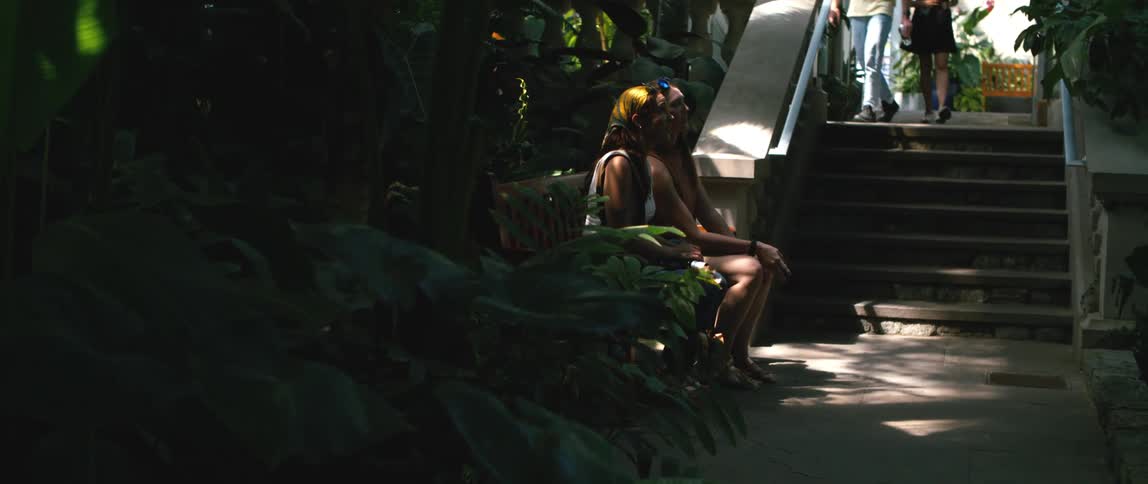 Two teenage girls are sitting on a bench in an interior garden while talking to each other and one of them points