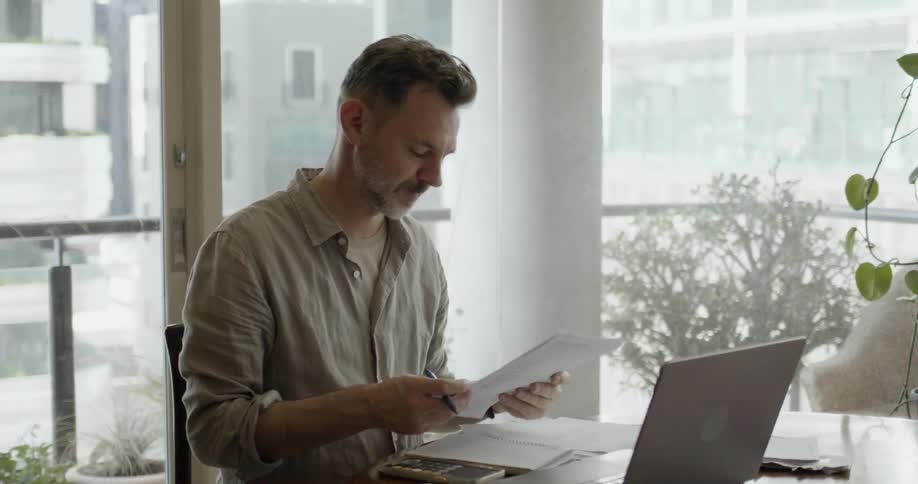 A man resting his face on his hand as he does math on a calculator and a laptop then looks at a paper and takes notes on a notebook