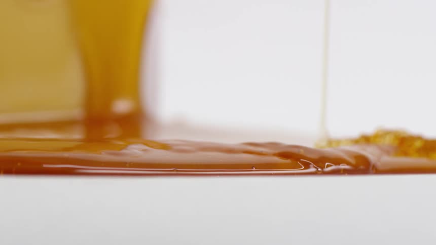 A line of honey drops down into an oozing puddle of more honey on a white surface