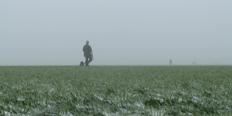 A man carrying a rifle walks in a field with his dog