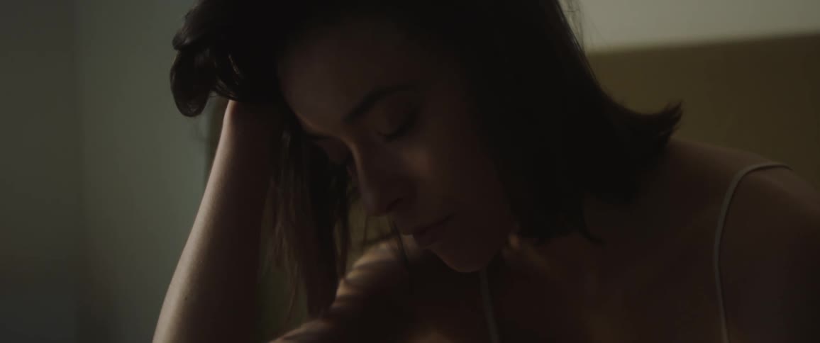 Sad woman in white undershirt sits on bed
