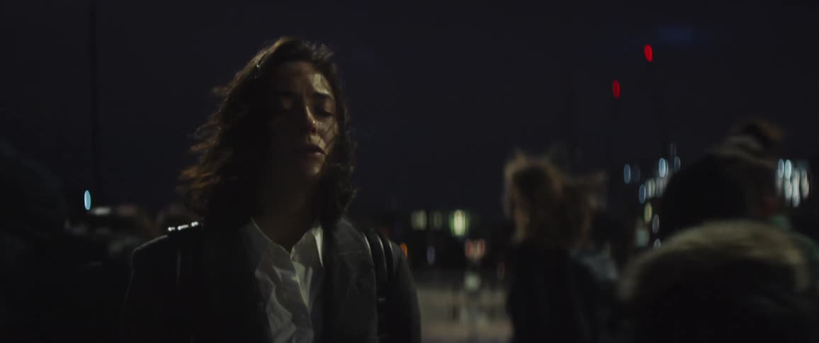 Woman stands crying with her eyes closed at night as pedestrians and vehicles move past