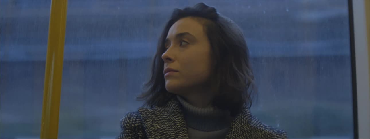 A woman is sitting on a train as it travels and she pushes her hair back with her hand