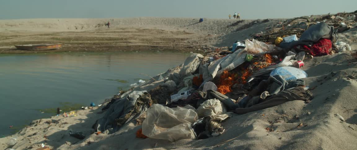 Garbage piled up in a heap in sand near water as people walk around in the far distance