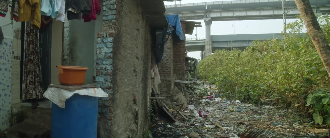 Clothes hang on a line outside a decrepit building with debris in puddles and freeway bridges in the distance