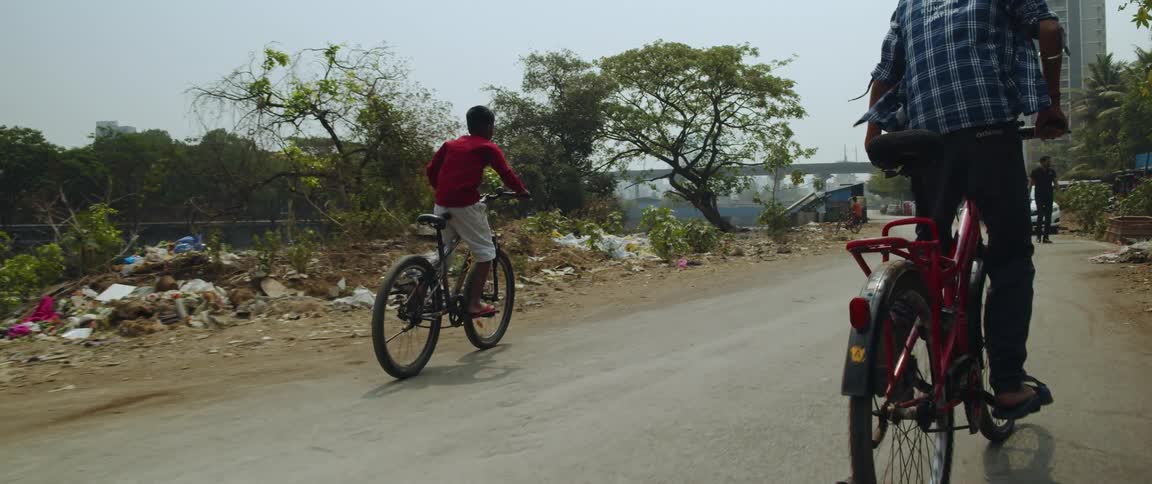 A teenager and a younger boy are riding bicycles on a road toward a tall building and a bridge as a man walks near a car