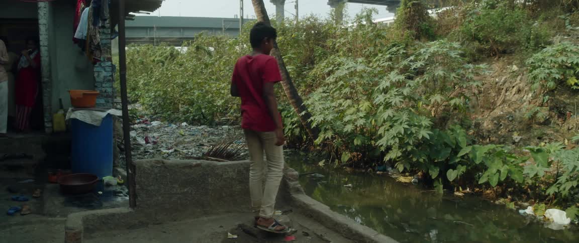 Boy with red t shirt and brown pants runs in alley to small river with litter