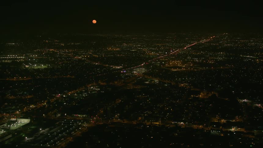 Steady traffic moves on the highway as lights glow throughout the city and an orange full moon shines in the night sky