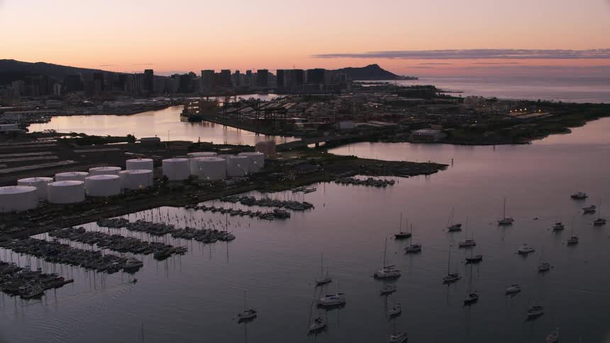 Bridge crosses mouth of harbor near storage tanks with city in distance under red sky